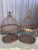 Pair of rustic metal birdcages shown outside on a stone surface against a corrugated metal backdrop. Both birdcages have domed tops with crown-shaped finials and vertical bars with ornamental metal banding.