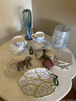 Overview of assorted items including Worchester bone china teacups and saucers, leaf dish, elephant brooch, decorative fruits, glass paperweights, and tile coasters on white table.