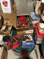 Box containing artificial red poinsettia flowers, red bows, faux greenery with pinecones, and boxes of string lights.