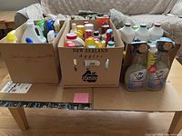 Three cardboard boxes filled with a variety of used cleaning supply bottles and jugs including spray bottles and gallon containers, some with colored triggers, some clear bottles of Zap brand cleaner on the side.