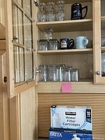 Cabinet shelves containing multiple glassware items including clear wine glasses, tumblers, and four ceramic mugs in various blues. Below the shelves are unopened boxes of Kirkland water filter cartridges (10-pack) and Brita filter cartridges (5-pack).