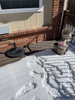 Wide shot showing the black metal umbrella stand laying on the ground, and the stacked concrete fountain positioned upright near a deck and a brick wall, partially covered in snow.