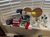 Photo showing multiple kitchen and houseware items arranged on a white cloth near a window with shutter blinds.
