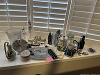 Wide view of all items including clock, glassware, figurines, and candles on tiled window ledge with white plantation shutters behind.