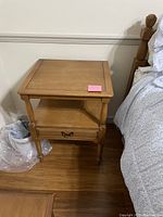 Front and side view of the wooden nightstand placed next to a bed on a hardwood floor. Shows drawer and shelf details.