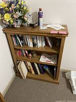 Oak bookcase with three shelves filled with books and decorative items, viewed frontally. Top surface holding various small items and artificial flowers.