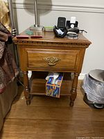 Oak nightstand showing front view with single drawer and lower shelf, mild surface wear, placed next to the bed and trash can.