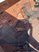 Two greenish-gray metal mesh patio chairs and a matching small metal mesh table arranged outdoors on stone patio.
