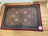 Top-down view of small rectangular rug with ornate traditional pattern, placed on hardwood floor in front of a door vent.