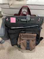 Stack of multiple travel bags including a large black bag and a brown Tumi branded bag at the front