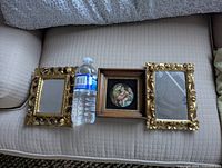 Top-down photo of the three small wall hangings: two gilded framed mirrors and one framed Madonna della Seggiola print, positioned on a couch with a water bottle for scale.