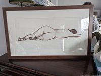 Photo of the framed artwork showing side view of the reclining nude woman in brown pastel, resting on a dark wooden surface.