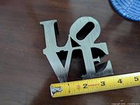 Front view of the brushed aluminum LOVE mini sculpture with a measuring tape showing width approximately 3.5 inches.