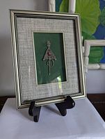 Close-up photo of the framed Moroccan Berber silver amulet on green velvet backing with textured gray mat and silver-tone frame, displayed on black stand.