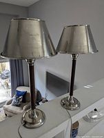 Photo of two identical Pottery Barn table lamps placed on a white ledge, showing their silver plated metal shades and bases, and mahogany stems.