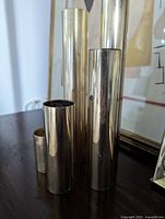 Five brass cylindrical tealight candle holders standing side by side on a dark wooden surface, showing varying heights and reflective brass finish.