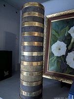 Tall cylindrical vintage brass vase with alternating smooth brass and dark ribbed bands next to framed floral art