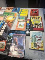 Assorted books and watercolor paint set on a table with visible titles including cookbooks and novels.