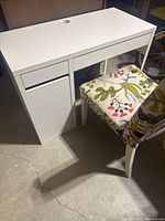 White desk and floral upholstered chair from front side, showing desk drawer and cabinet door.