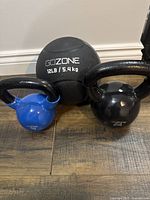Three GoZone fitness items on wood floor: blue 20 lb kettlebell, black 35 lb kettlebell, and black 12 lb weight ball.