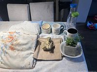 Photo showing folded floral and checkered bed sheets, three ceramic mugs with printed text, and two glass juice containers with blue and green lids, plus decorative potted plants on a table.
