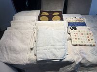 Photo showing folded white towels with a textured pattern and stacked pillow cases including brown with yellow circles and others with colorful designs.