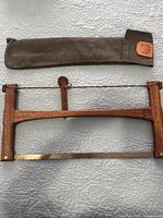 Photo of wooden outdoor folding saw with its brown protective fabric case on a textured surface.