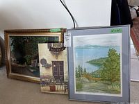 Two framed prints leaning against a wall, one large scenic print and a smaller artwork with patio scene; the main focus is the large print with a metal decor piece on it.