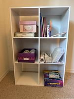 White bookshelf with six compartments containing books, storage boxes, paper rolls, plastic sleeves, and a board game.