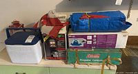 Photo of camping supplies stacked on a surface showing a blue cooler, Coleman propane lantern box, a tent box, green Coleman camp stove box, and a blue tarp.