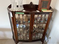 Front view of wooden curio cabinet showing glass front doors and decorative carved wooden details on top and legs, with various collectible items inside (which are not included in sale).