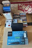 Overall lot view with stacked DVDs in cases, black storage case, and boxed Sony Blu-ray Disc/DVD player on carpet.