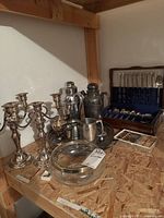 Photo showing four silver-tone candlesticks, two cocktail shakers, a silver-tone pitcher, a matching tray, a large serving bowl, a boxed flatware set and a set of knives with mother-of-pearl handles.
