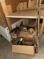 View of cardboard boxes containing fishing tackle boxes, fishing accessories, and a straw hat nearby under wood shelf in basement.