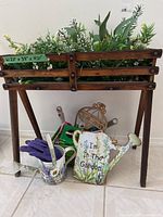 Small wooden plant stand filled with artificial plants, decorative watering can, garden sign, and gardening gloves displayed.