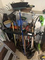 Assortment of garden hand tools including rakes, brooms, pruners, garden fork, and boots arranged on a rack and floor.