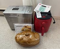 Three items grouped on carpet: Breadman bread maker, Dash air fryer, and ceramic serving dish. Bread maker is stainless steel with digital controls, air fryer is red with black handle, serving dish is brown ceramic with lid.