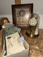 Photo showing partially constructed doll in box with paper doll outfits, the framed coin collection, and mantelpiece clock under glass dome.