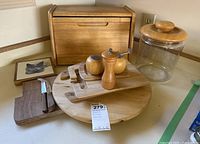 Wooden kitchenware items displayed including bread box, round platter, cutting boards, pepper grinder, salt shaker, and glass jar with wooden lid.