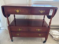 Front view of wooden drop-leaf table showing turned legs, drop leaves on both ends, five drawers with brass handles and caster wheels