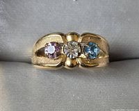 Front view of ring showing three round faceted stones: purple, clear, and blue, set in a heavy 14K gold plated band with textured detailing.