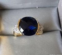 Photo showing the ring face with large round dark blue faceted stone and clear accent stones on the sides.
