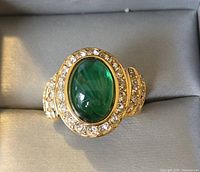 Close-up front view of gold-tone ring with oval green stone and surrounding clear rhinestones.