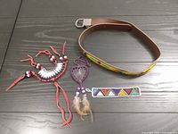 Full view of the lot showing the beaded leather belt, the red leather and bead necklace, the purple heart-shaped dreamcatcher necklace with feathers, and the beaded bracelet on a black wooden surface.