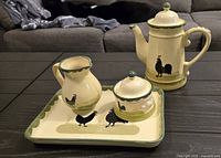 Photo of the complete ceramic tea set on a dark wood table showing teapot, creamer, sugar bowl, and tray with rooster designs and green scalloped edges.