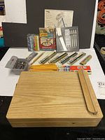 Wooden travel art box with easel lid and assortment of art supplies including pens, colored pencils, pastels, charcoal, and erasers arranged on table