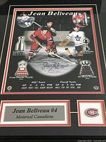 Close-up of framed photo showing Jean Beliveau in Canadiens red jersey during faceoff against Toronto Maple Leafs player, with trophies and stats. Also visible is Jean Beliveau's signature.