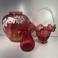 Lot showing all three cranberry glass items together: large hand painted floral vase, smaller ruffled rim vase, and cranberry glass basket with clear handle.