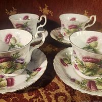 Four Royal Albert Highland Thistle teacups and matching saucers arranged on patterned fabric, showing floral design and gold trim