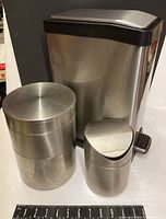 Three stainless steel accessories shown together including a large rectangular foot pedal waste can, a small oval swing top waste can, and a three-level stacked storage drum.